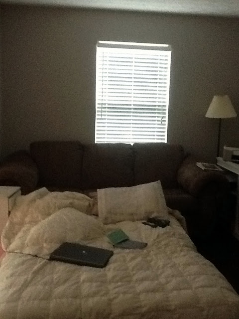 Dim interior photo of a bedroom with a quilted bed, brown couch, lamp, and window with blinds letting in light; muted browns, low-key warm tones, soft shadows, calm and understated mood.